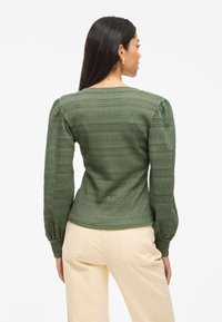 Pull en maille verte avec des manches bouffantes, motif texturé de rayures horizontales et une silhouette légèrement ajustée. Associé à un pantalon beige clair.