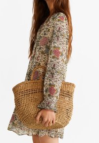 Sac panier en paille tissée avec poignée circulaire, présentant un design en tressage ouvert. Tons neutres avec une texture naturelle, porté avec une robe à motif floral.