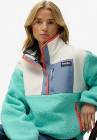 Pull en polaire de couleur vert menthe et crème, avec un col haut, une poche poitrine zippée, et des accents orange et bleu contrastants.