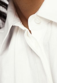 Chemise blanche à boutons avec un col pointu et un bouton visible au cou. Le tissu semble lisse et légèrement texturé.