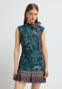 Kurzärmliges grünes Kleid mit Paisleymuster, Schleifen-Detail am Kragen, strukturiertem Stoff und gerafftem Saum in kontrastierenden lila und braunen Akzenten.