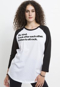 Biała koszulka raglanowa z czarnymi rękawami, ozdobiona wyraźnym czarnym tekstem: "Bądź miły. Troszczcie się o siebie nawzajem. Słuchajcie alt rocka." Gładka faktura.