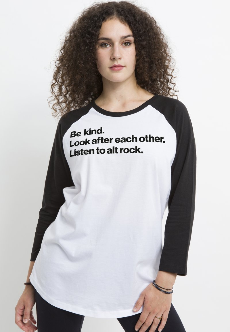 Vit raglan-t-shirt med svarta ärmar, med fet svart text: "Var snäll. Ta hand om varandra. Lyssna på alt rock." Slät textur.