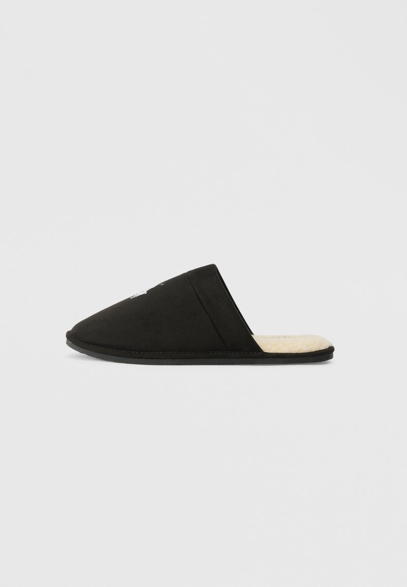 Schwarzer Slip-On-Schuh mit geschlossenem Zehenbereich und cremefarbenem, weichem Innenfutter, von der Seite auf einem schlichten, hellen Hintergrund betrachtet.