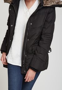 Svart pufferjacka med huva i päls, dragkedja framtill och sidofickor. Materialet verkar vara vävt med en jämn yta.