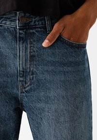 Mörkblå denimjeans med synliga sömmar, femfickermodell och knappstängning. Närbild som fokuserar på midjebandet och handen i fickan.