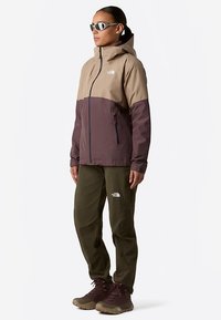 Brun och beige vattentät jacka med huva, dragkedjefickor, kombinerad med gröna byxor och bruna vandringskängor. Mjuk textur, utomhusdesign.