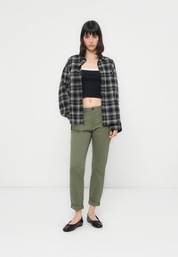 Kvinne som står mot en hvit vegg iført en svart og hvit rutete skjorte over en svart crop top, olivengrønne bukser og svarte leppestift.