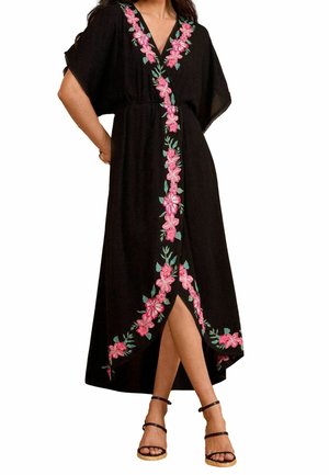 Mujer con vestido midi negro de escote en V con bordados florales rosas a lo largo del escote y el dobladillo, combinado con sandalias negras de tiras en el tobillo.