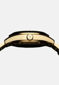 Goldene Uhr mit einem schwarzen strukturierten Lünette und einem kreisförmigen Logo an der Seite. Verfügt über eine glatte metallische Oberfläche und ein filigranes Design am Armband.