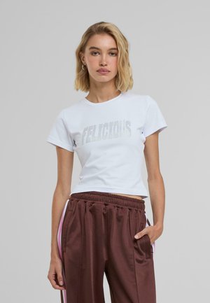 Fehér pamut póló "FELICIOUS" ezüst betűkkel, barna, rugalmas derékú jogger nadrággal, pink oldalsávokkal párosítva.