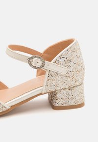 Sandalia blanca con brillo y tacón bloque, con hebilla plateada y correa en el tobillo. La parte superior tiene una textura brillante y un forro liso.