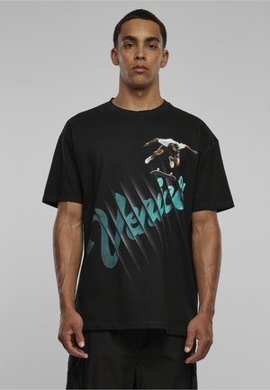 Homme portant un t-shirt noir avec un texte turquoise « Venice » et un graphisme d’un skateboarder réalisant un saut sur un fond gris.