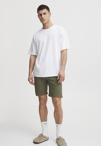 Hvit bomulls T-skjorte med løs passform, kombinert med grønne shorts. Modellen har også på seg hvite sokker og brune slip-on sko. Nøytral bakgrunn.