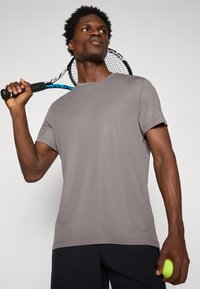 Grå atletisk t-shirt med en tekstureret overflade, korte ærmer og logo-detalje på ærmet, båret af en person der holder en tennisketsjer og bold.