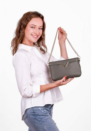 Olivgrüne Lederhandtasche mit einem goldfarbenen V-förmigen Akzent, Kettenriemen und Reißverschluss. Das Modell hält sie vor einem weißen Hintergrund.