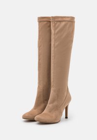 Une paire de bottes en daim beige montantes avec des pointes et des talons aiguilles sur un fond blanc.