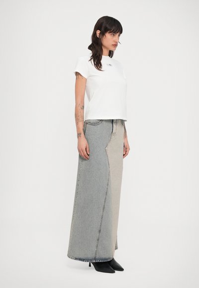 Nő, fehér pólóban, kétszínű hosszú farmer szoknyában és fekete, hegyes orrú, magassarkú csizmában áll, egyszerű fehér háttér előtt.