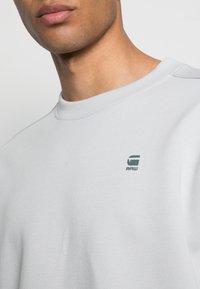 Sweatshirt gris clair avec un col rond, texture douce et un petit logo vert "RAW" sur la poitrine gauche. Design minimaliste.