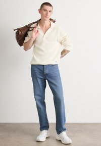 Kremfarget strikkegenser med myk tekstur, blå jeans og hvite sneakers, holder en brun lærveske over skulderen.