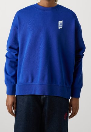 Persona con una sudadera azul brillante con un logo blanco en el pecho y vaqueros azul oscuro con un diseño bordado en rojo.