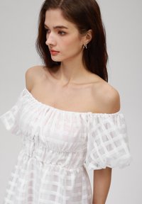 Robe blanche à épaules dénudées avec un motif à carreaux, en tissu transparent, corsage froncé et manches bouffantes. Présente une coupe décontractée et une texture douce.