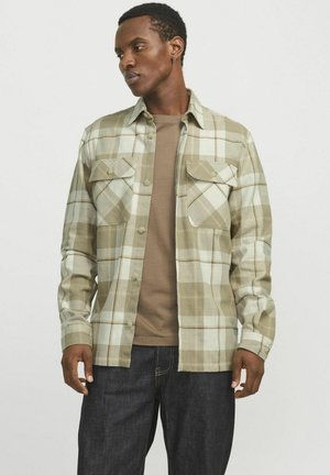 Plaid long-sleeve shirt in light brown and green tones, featuring two chest pockets and a button-up front. Paired with a brown T-shirt.