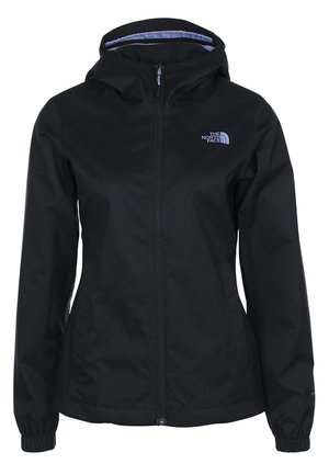 Frau trägt schwarze Kapuzenjacke, bordeauxrotes Shirt, beige Cargohose und braune Wanderschuhe und steht vor einem hellgrauen Hintergrund.