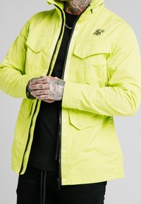 Neon gul jakkesæt med lynlås foran, to brystlommer og broderet logo på venstre side. Tekstureret stof, lange ærmer, tætsiddende design.