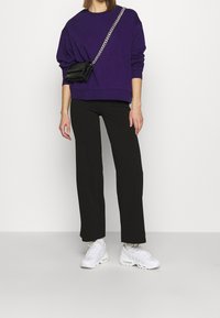 Sudadera oversized morada, pantalones de pierna ancha negros, zapatillas blancas y un pequeño bolso cruzado negro con correa de cadena.