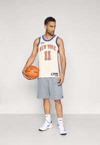 Basketballspieler in einem cremefarbenen Trikot mit blauen und orangenen Akzenten, Nummer 11, graue Shorts, hält einen Basketball und trägt weiße Turnschuhe.