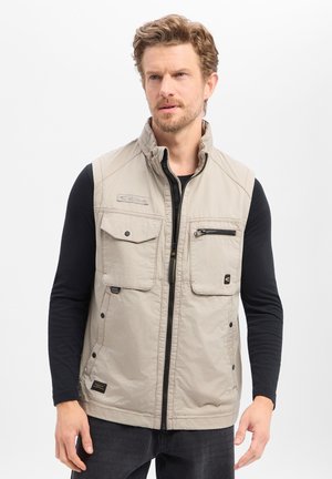 Man draagt een beige mouwloos vest over een zwart shirt met lange mouwen, staand tegen een effen witte achtergrond.
