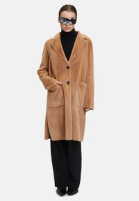 Cappotto in ecopelliccia beige con revers a intaglio, due tasche frontali e due bottoni, abbinato a un dolcevita nero e pantaloni a gamba larga.