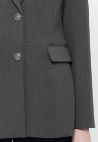 Blazer grigio con taglio sartoriale, dotato di due bottoni argentati, una trama sottile e una piccola tasca frontale. Linee pulite e design strutturato.