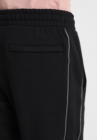 Schwarze Sweatpants aus weichem Material, mit lockerem Schnitt, elastischem Bund und einem grauen Seitestreifen. Verfügt über eine Paspeltasche auf der Rückseite.