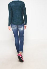 Ciemnozielony sweter dzianinowy, dopasowane niebieskie dżinsy i różowe sneakersy. Długie rękawy, okrągły dekolt i lekko zaokrąglony tył.