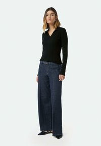 Schwarzes, figurbetontes, geripptes Langarmoberteil kombiniert mit hochgeschnittenen, weit geschnittenen Jeans aus dunklem Denim, abgerundet mit kontrastierenden gelben Nähten und spitzen schwarzen Schuhen.