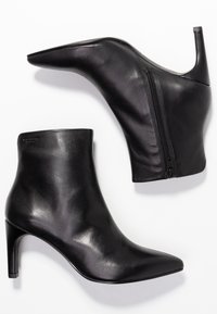 Botines de tacón alto negros, hechos de cuero suave, con punta afilada y cremallera lateral para un fácil calzado. Detalles de botón en el tacón.