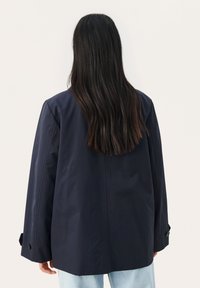 Mørkeblå oversized jakke med en glat tekstur, stort rygudskæringsdesign og knapdetaljer på ærmerne, parret med lyseblå jeans.