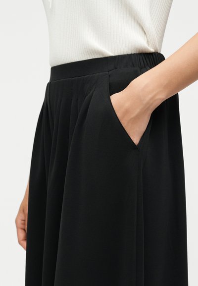 Άτομο που φοράει μαύρη πιετέ φούστα με ελαστική μέση, χέρι τοποθετημένο στην πλαϊνή τσέπη, συνδυασμένη με λευκό ριμπ τοπ.