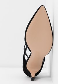 Chaussure à talon haut noire avec bout pointu, dessus en daim texturé, brides latérales transparentes et semelle couleur nude.