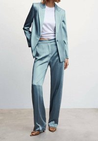 Costume en satin bleu clair avec un blazer structuré et un pantalon ample, associé à un haut court blanc. Texture lisse et lignes nettes.