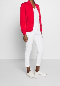 Röd bomberjacka med en slät, glänsande yta, dragkedja fram och ribbade manschetter. Stylistiskt kombinerad med figurformande vita jeans och vita sneakers.