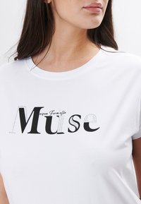 Hvid bomulds t-shirt med fed sort tekst "Muse" med rhinsten, rund halsudskæring og korte ærmer.