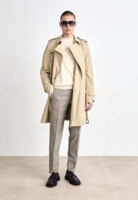 Beige trench coat vállpántokkal, világos krémszínű pulóver és világos szürke kockás nadrág. Sötétbarna, vastag talpú cipők, fekete napszemüveg.