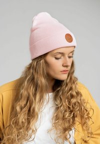 Różowy, prążkowany czapka beanie z małą, okrągłą brązową skórzaną łatką z przodu, noszona w luźnym stylu z żółtą kurtką na białej koszuli.
