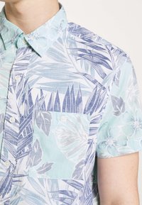 Koszula na krótki rękaw, zapinana na guziki, w jasnoniebieskim kolorze z kwiatowym i liściastym wzorem w odcieniach niebieskiego i szarości, z kieszenią na piersi.