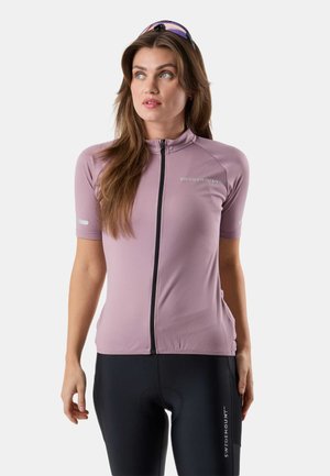 Vrouw die een lichtpaarse fietsjack met ritssluiting en zwarte fietsshorts draagt, lichtjes naar links kijkt en een zonnebril op haar hoofd heeft.