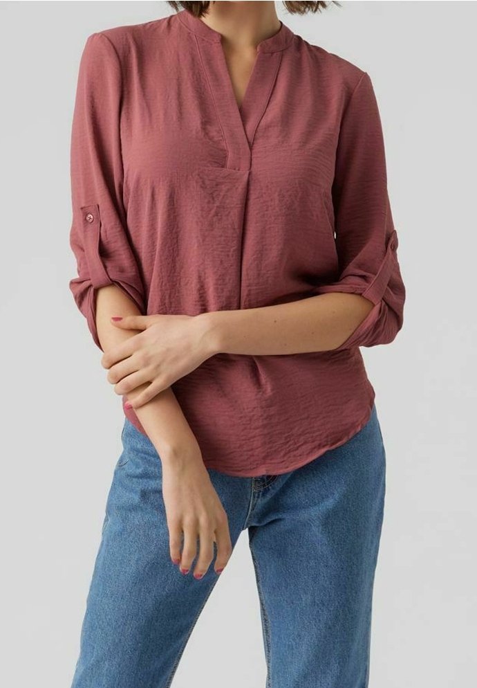 Blusa rosa con scollo a V e maniche arrotolate, realizzata in tessuto texture. Indossata con jeans blu, che mostrano una vestibilità rilassata.