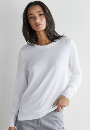 Frau mit mittellangen dunklen Haaren, die ein schlichtes weißes Langarmshirt und hellgraue Hosen trägt, steht vor einer weißen Wand.